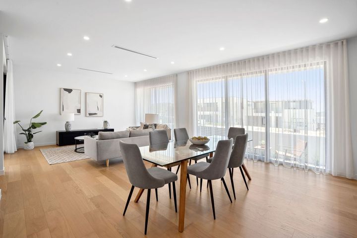 Bright open-plan living and dining room with wood floors, gray chairs, and large windows with sheer curtains