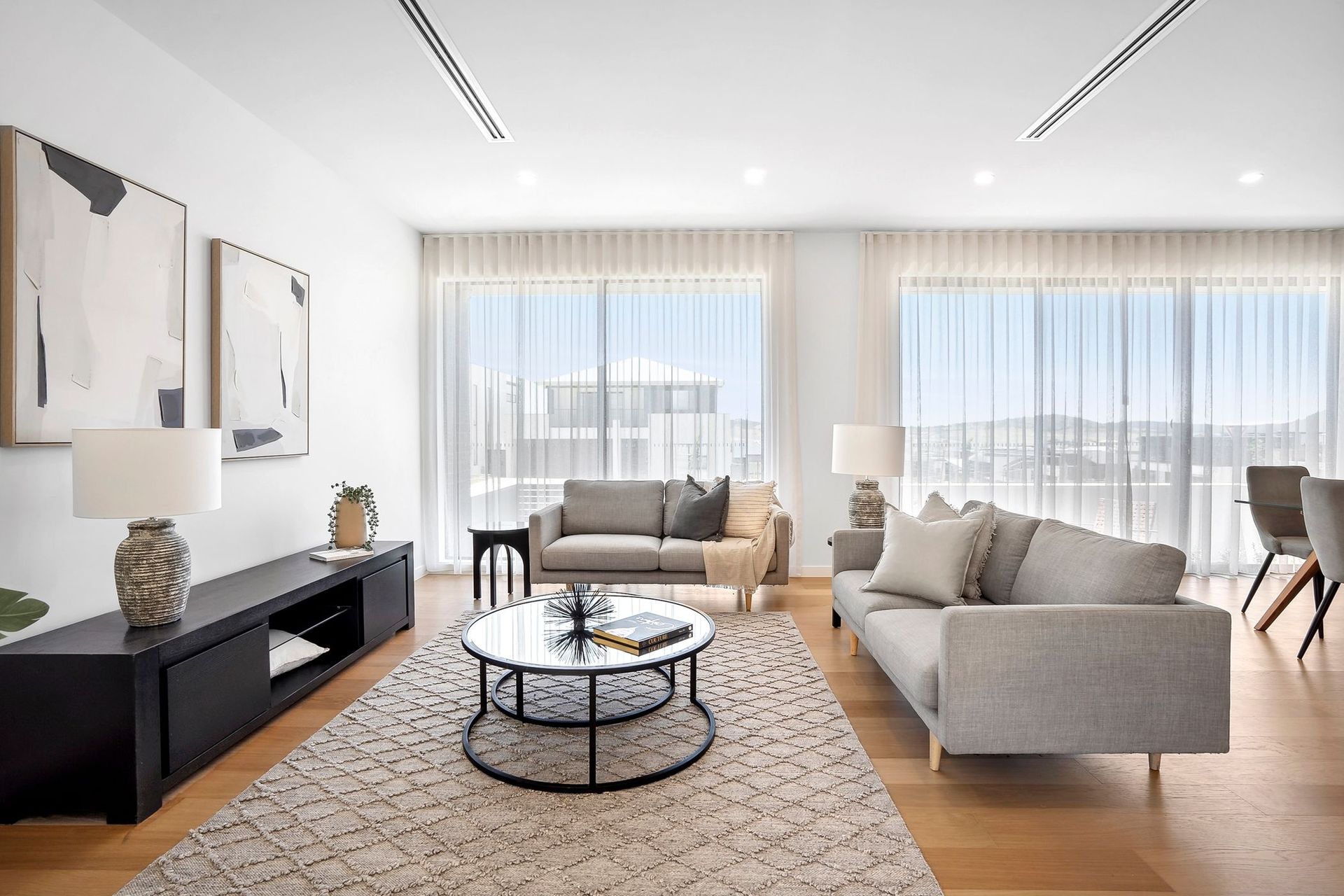 Bright modern living room with beige sofas, round coffee table, rug, TV console, and large sunlit windows