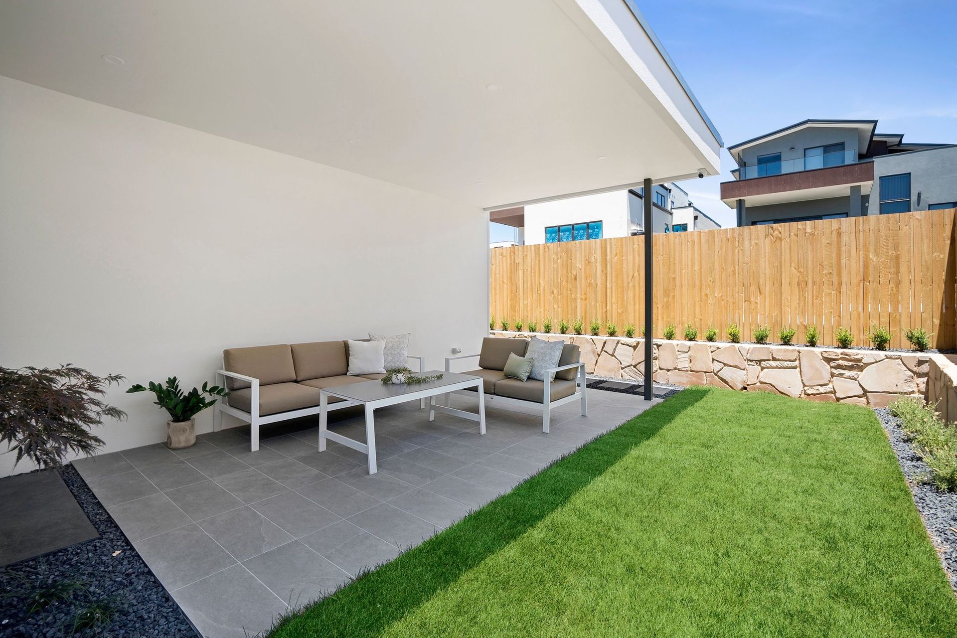 Covered patio with gray outdoor furniture, green lawn, and wooden fence in a modern backyard.