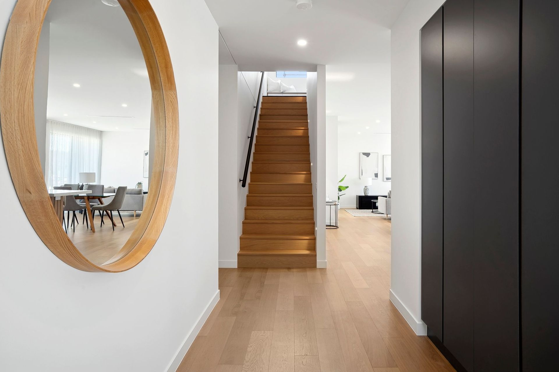 Modern hallway with wooden stairs, black cabinet, round mirror, and light wood flooring