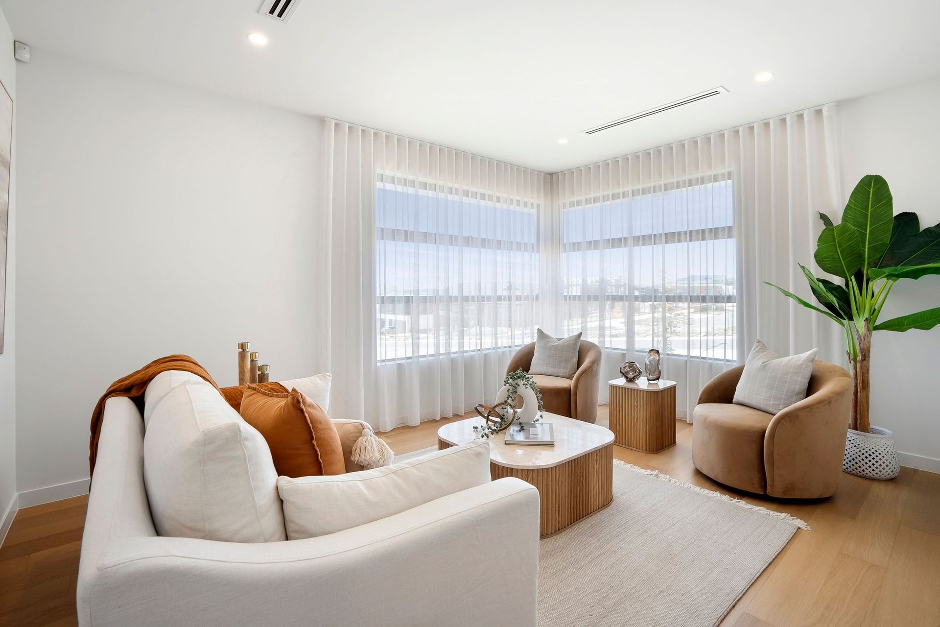 Bright modern living room with white sofa, tan chairs, round wood tables, and large windows.