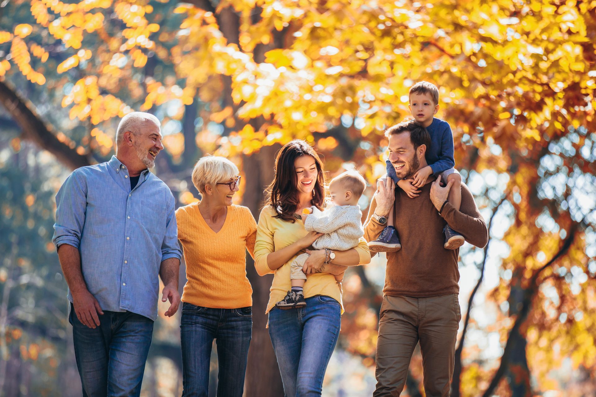 Family walking outdoors in autumn, smiling among orange leaves, with a child on the father's shoulders.