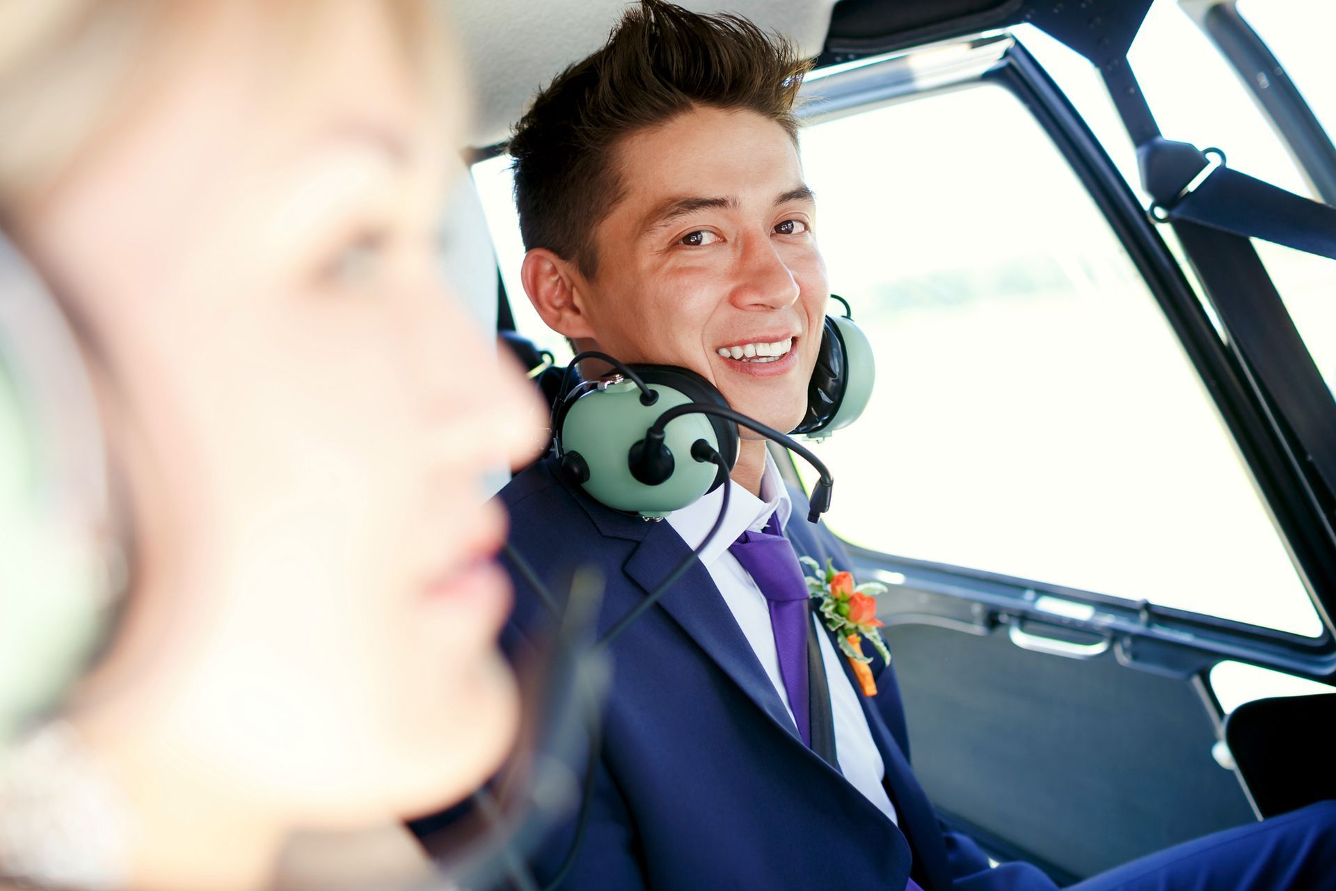 Smiling pilot in cockpit wearing headset, with a blurred passenger in the foreground
