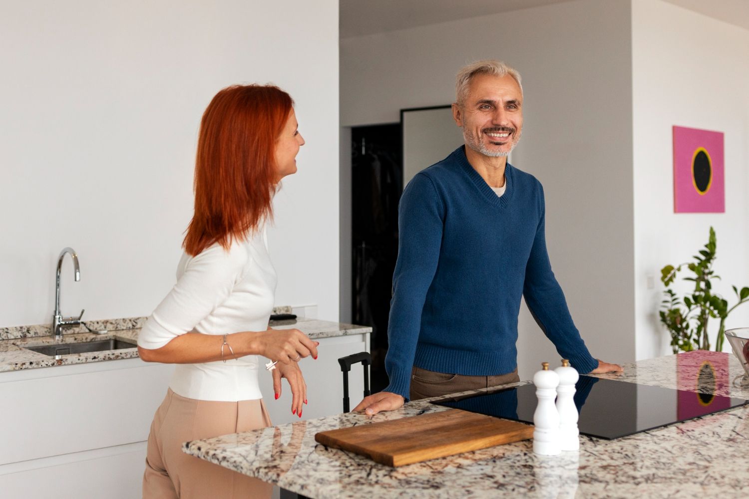 Woman and man chatting in a bright kitchen beside a granite island and cutting board.
