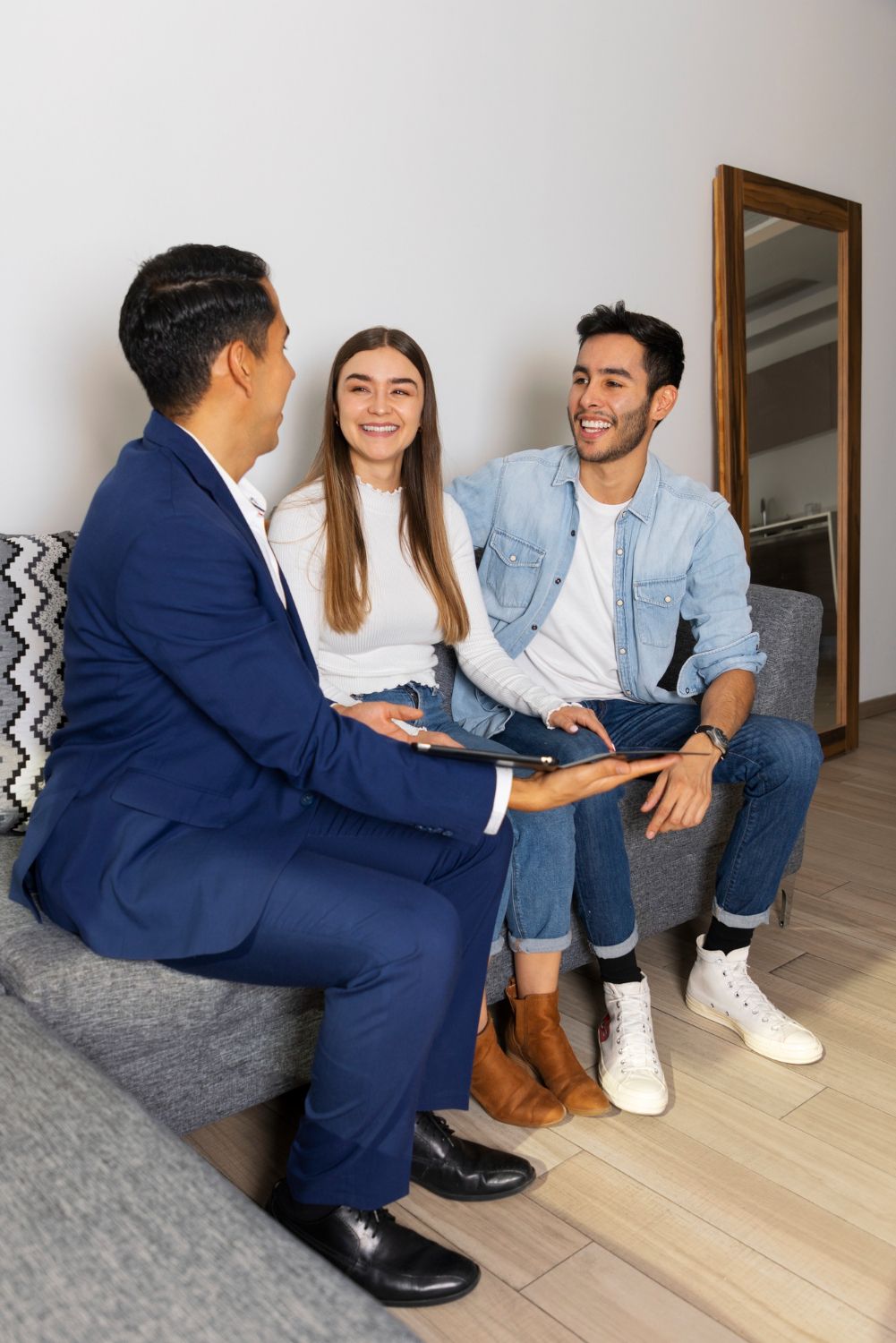 Three people sit talking on a sofa in a bright living room, with a mirror and wood floor nearby.