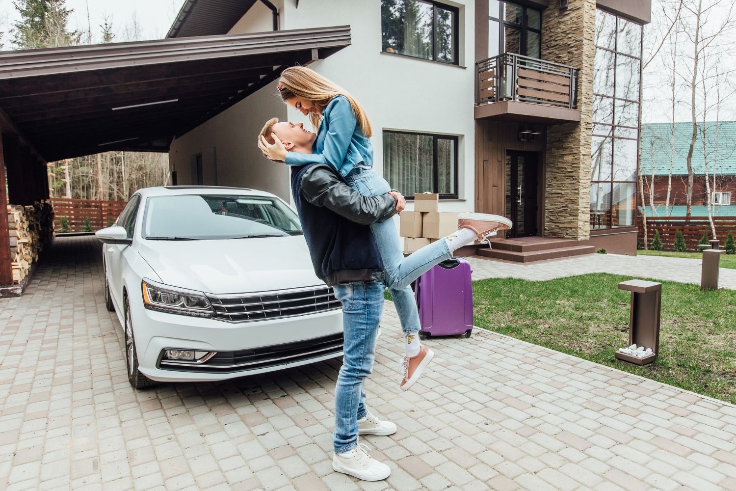 Two people embracing beside a white car in a driveway, with a purple suitcase nearby.