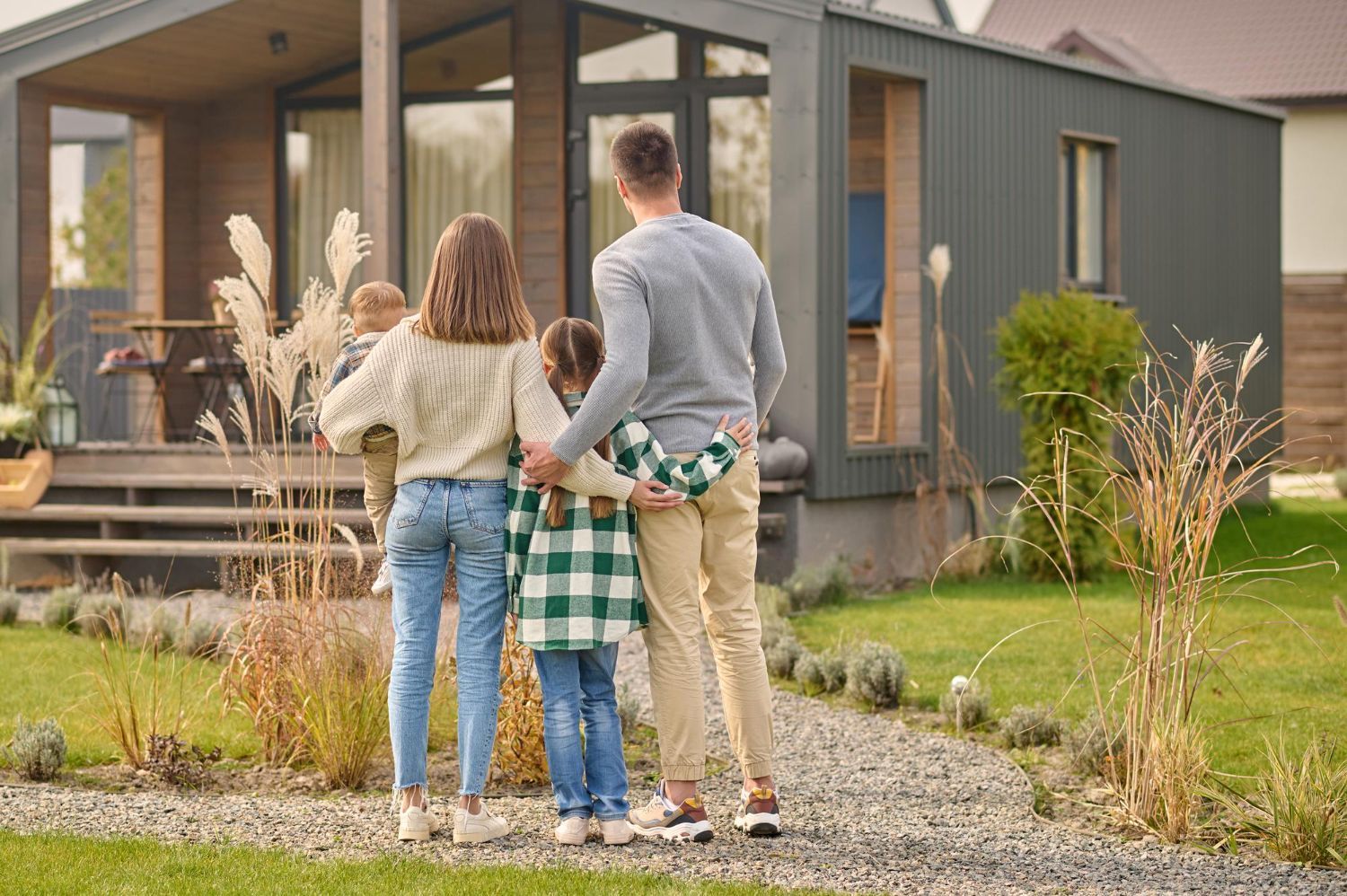 Three people stand on a path, facing a modern gray house with a porch and large windows.