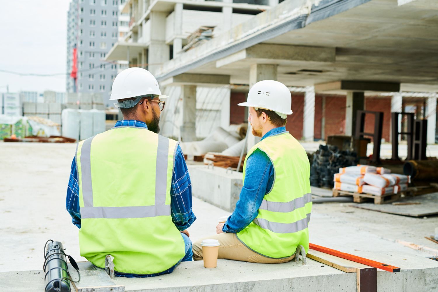 Two construction workers in hard hats and neon vests sit at a building site, facing a concrete structure.