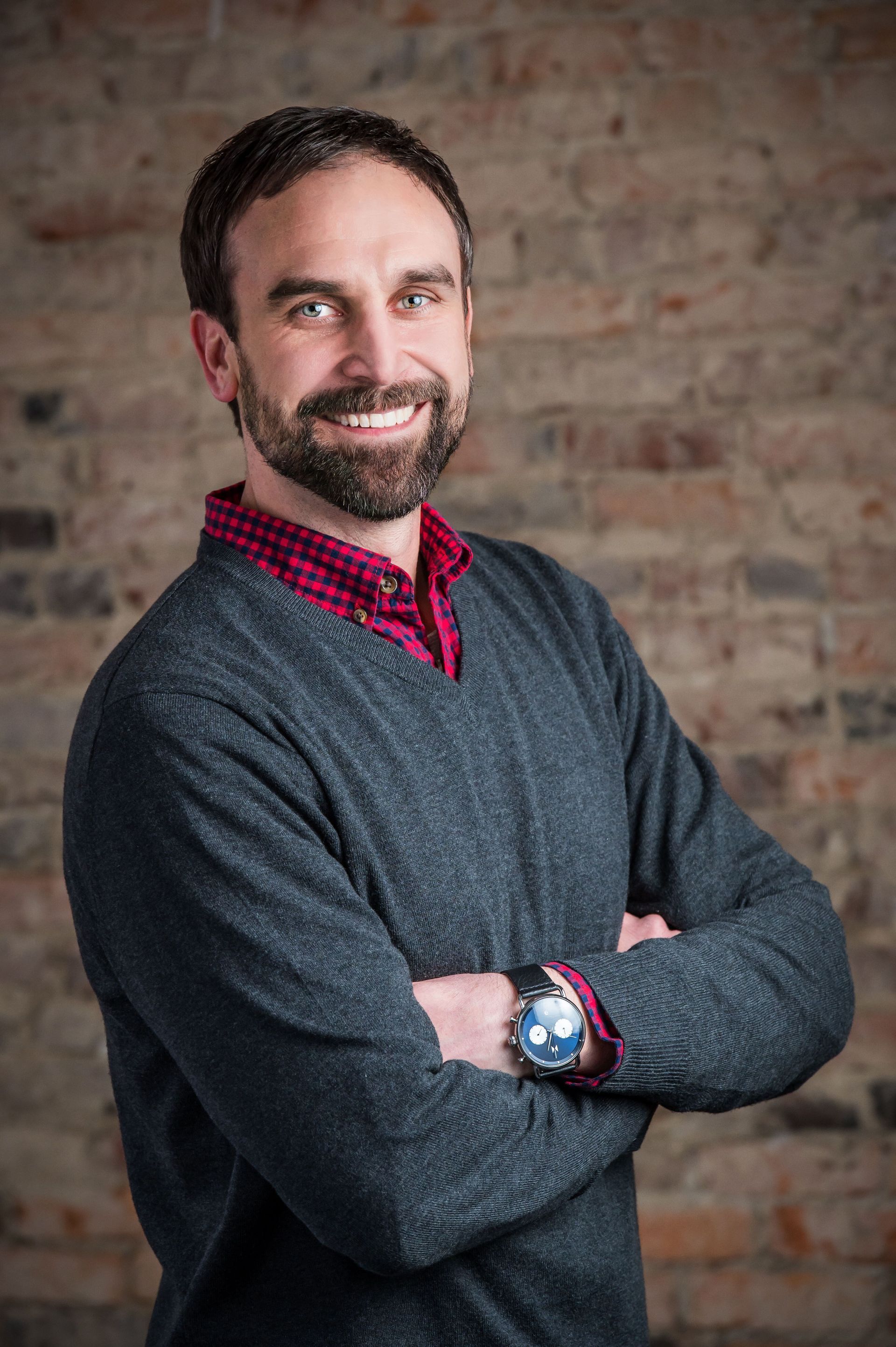 Smiling man with folded arms in a gray sweater against a brick wall background
