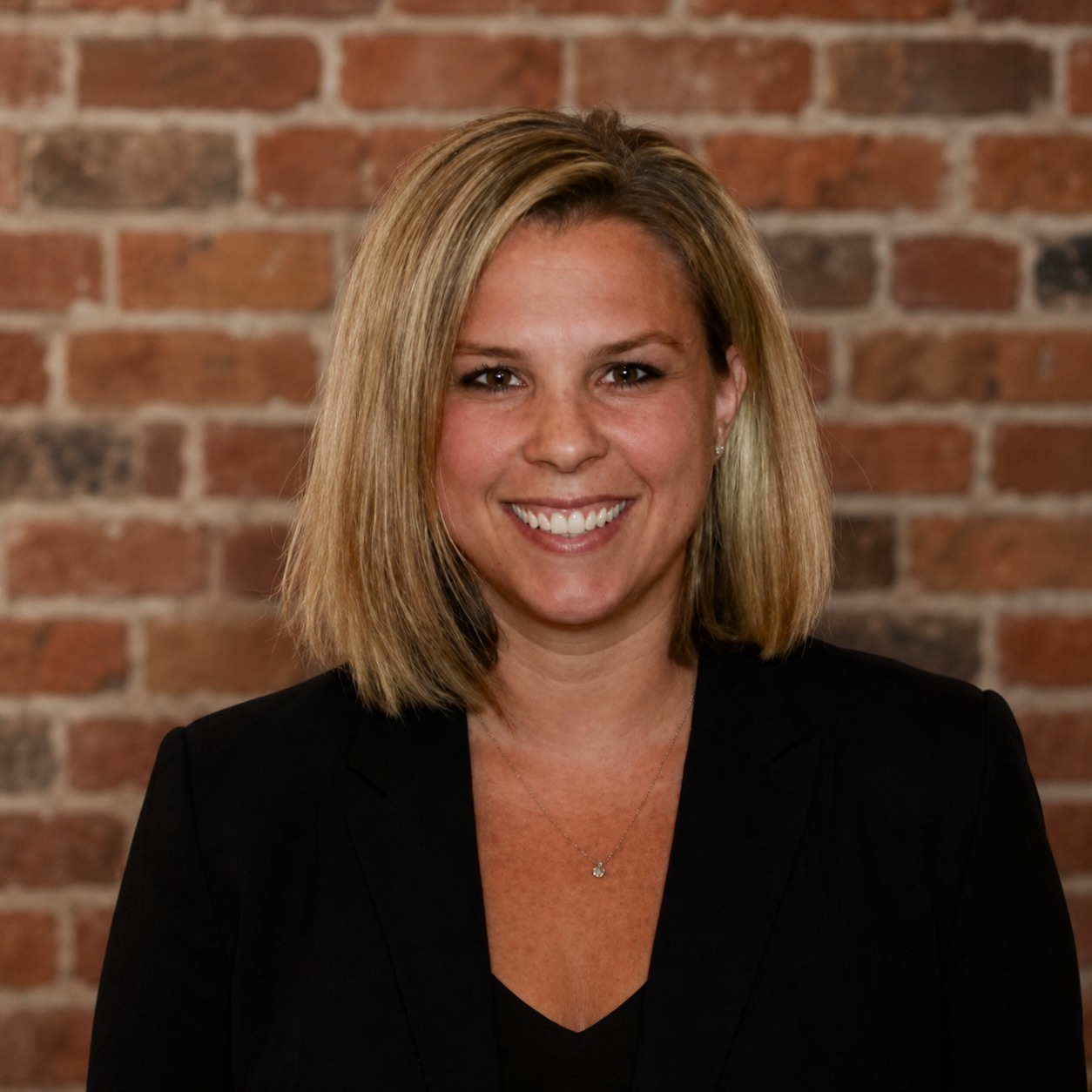 Smiling person in black blazer against a brick wall backdrop