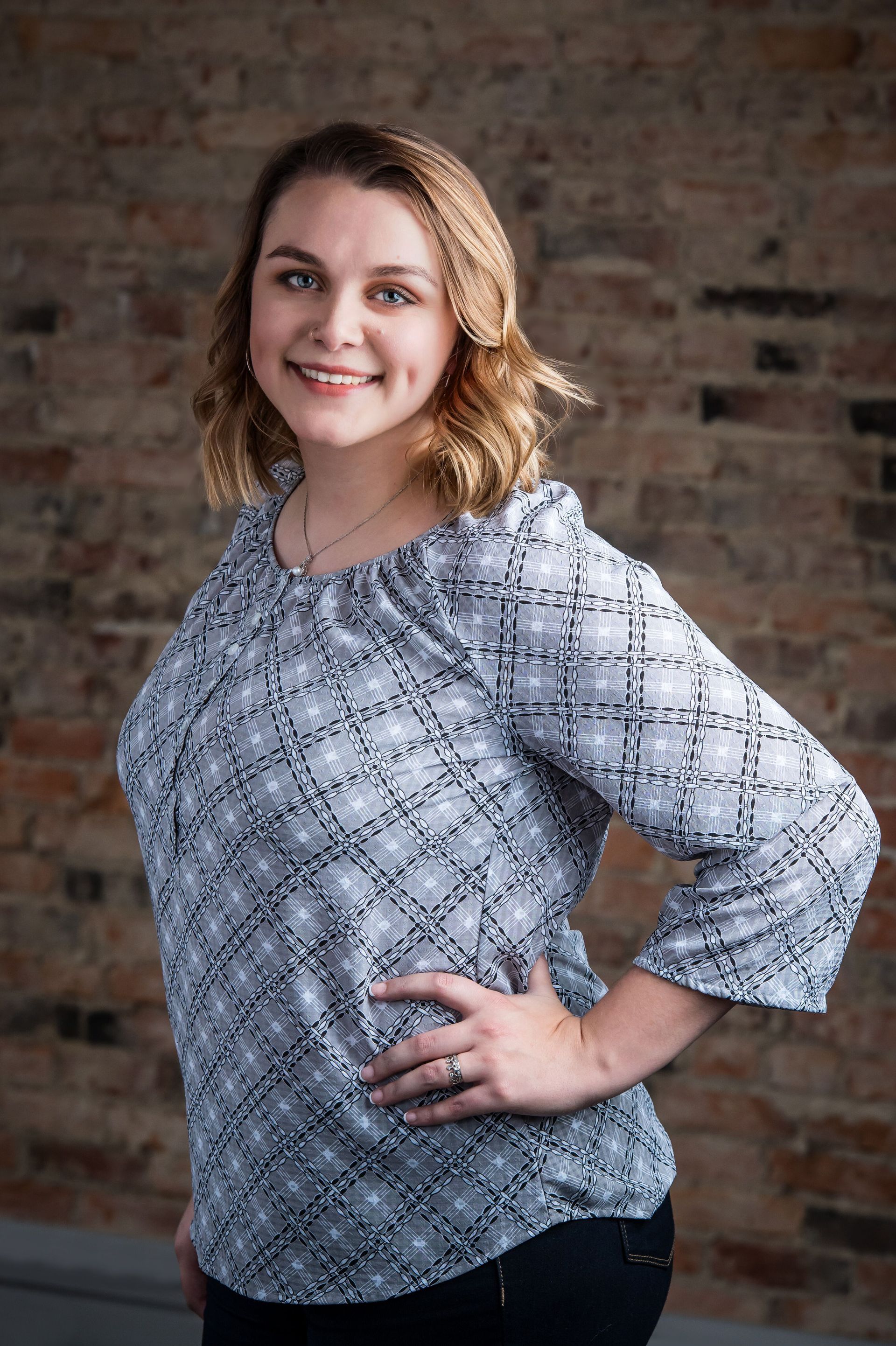 Smiling woman in a patterned blouse posing with one hand on her hip against a brick wall.