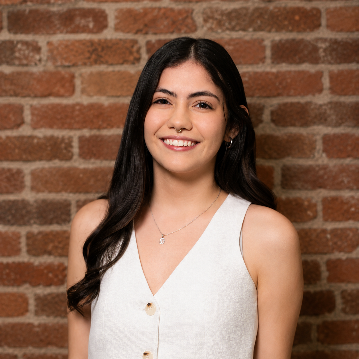 Smiling person in a white sleeveless top standing against a brick wall.