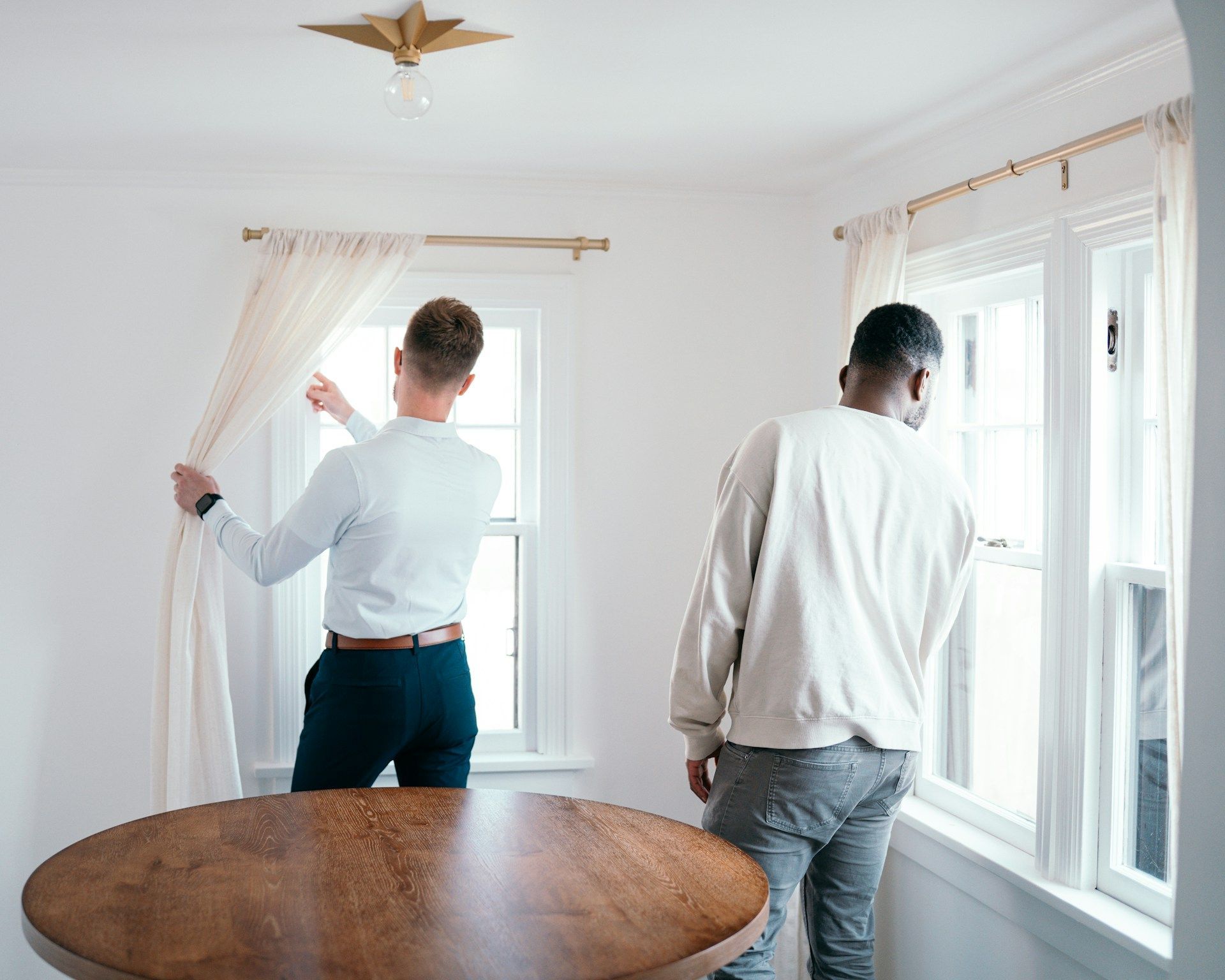 Two people opening white curtains in a bright room with large windows and a round wooden table