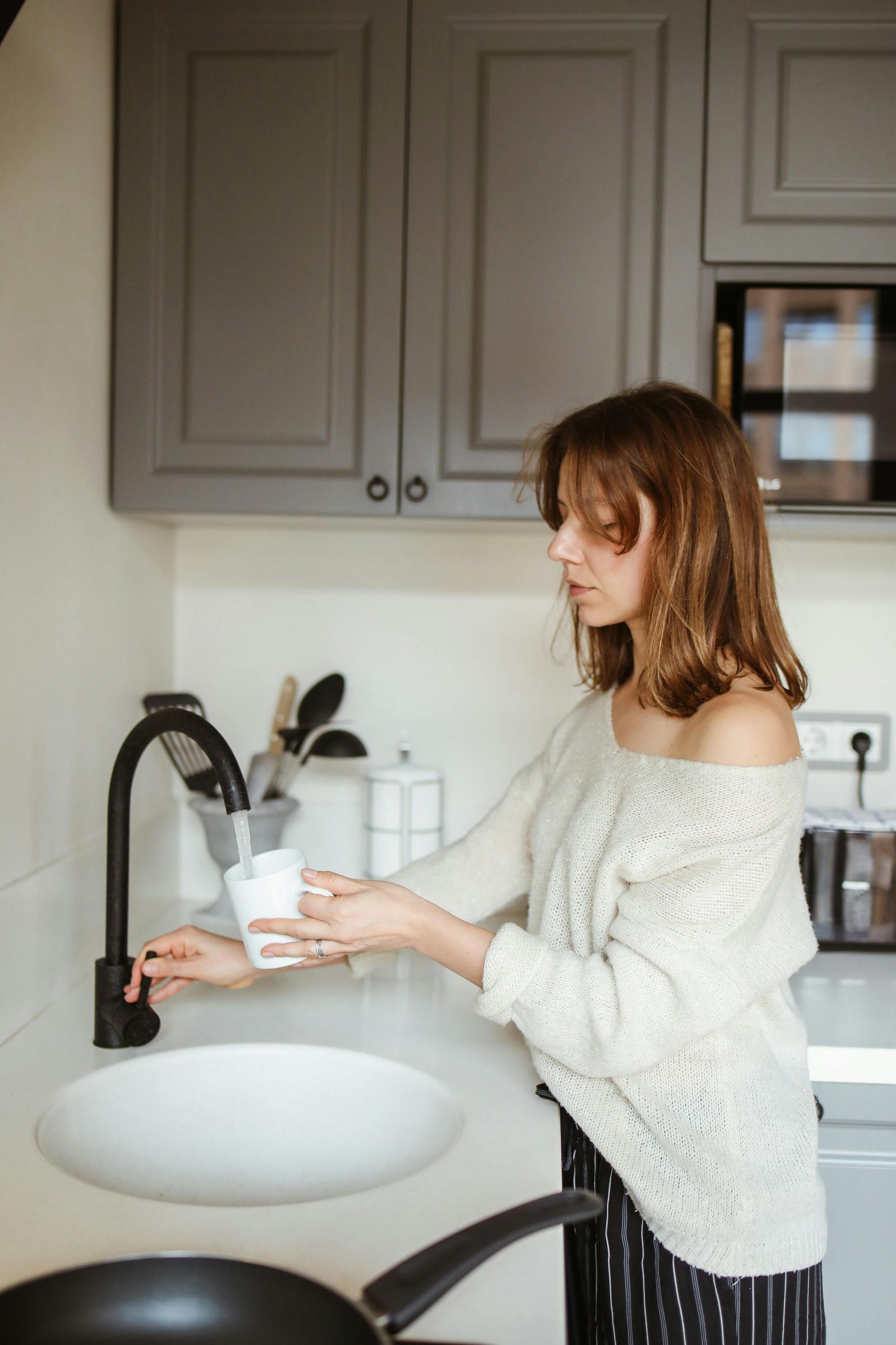 Woman filling a glass at a kitchen sink with gray cabinets and a black faucet