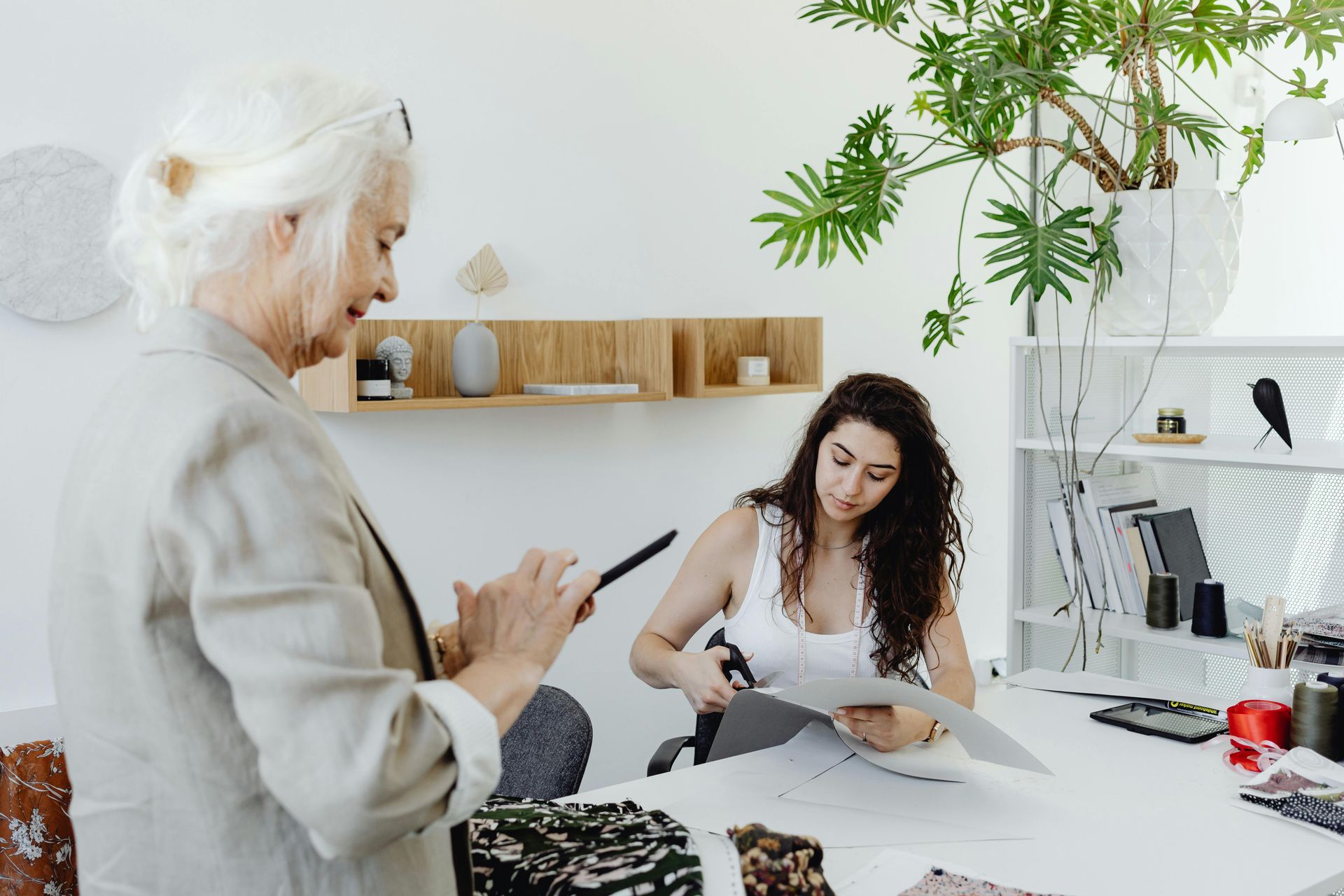 Two women in a bright studio, one sketching while the other reviews a tablet near a desk and computer.