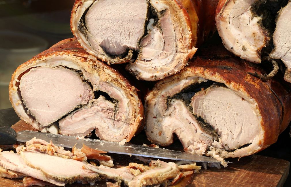 Primo piano di involtini di porchetta arrosto, con un coltello che affetta la carne su un tagliere di legno.