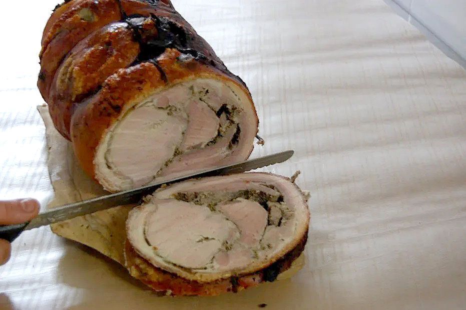 Una mano taglia una fetta da un arrosto di maiale arrotolato, ripieno di erbe aromatiche e di colore dorato, su una superficie bianca.