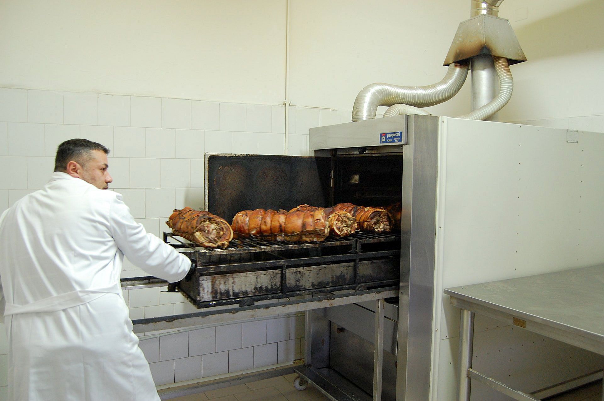 Una persona in camice bianco fa scivolare una griglia di carne arrosto in un grande forno industriale in una cucina professionale.