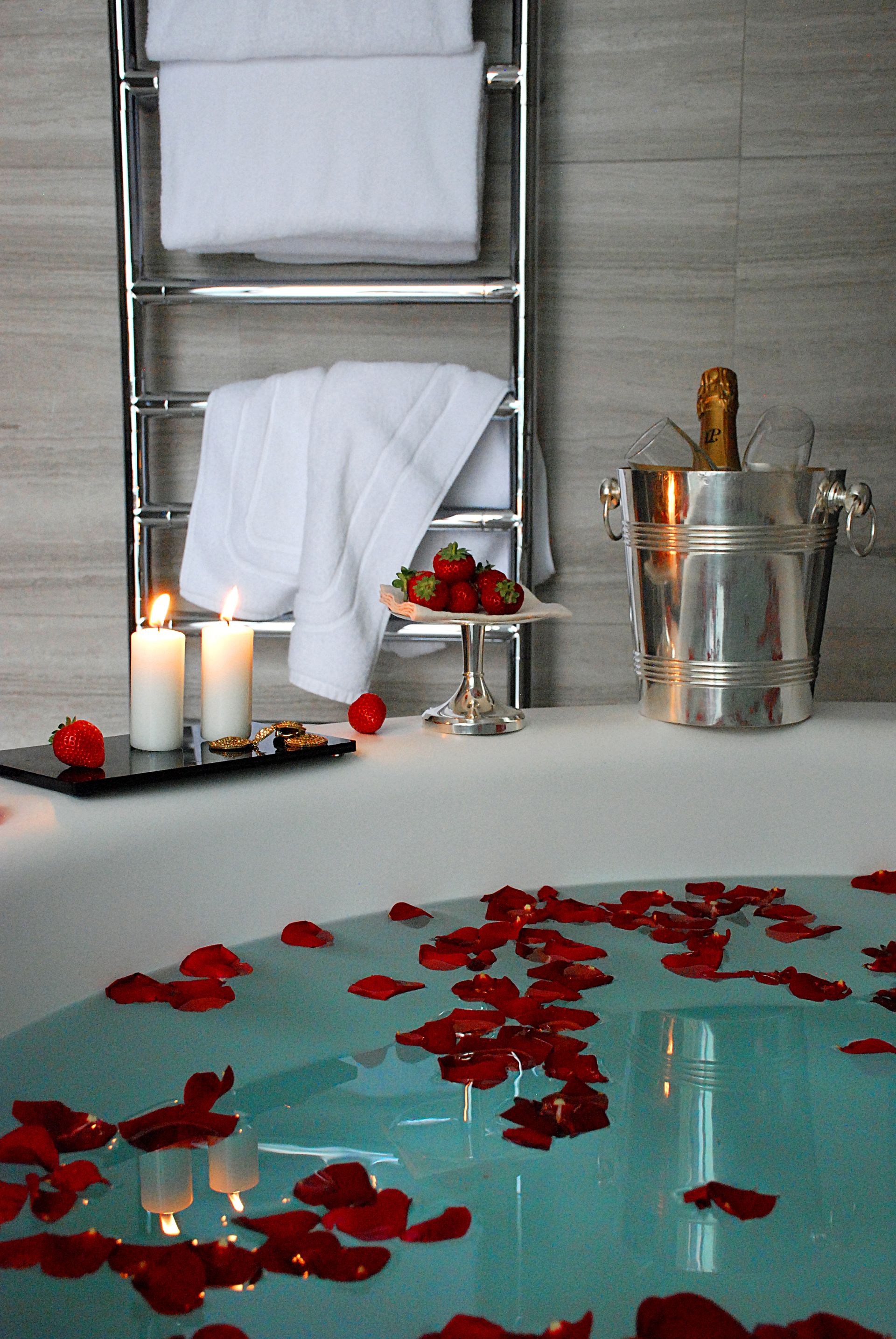 Ванна, наполненная водой и лепестками роз, рядом свечи, свежая клубника и ведерко для шампанского на подставке.