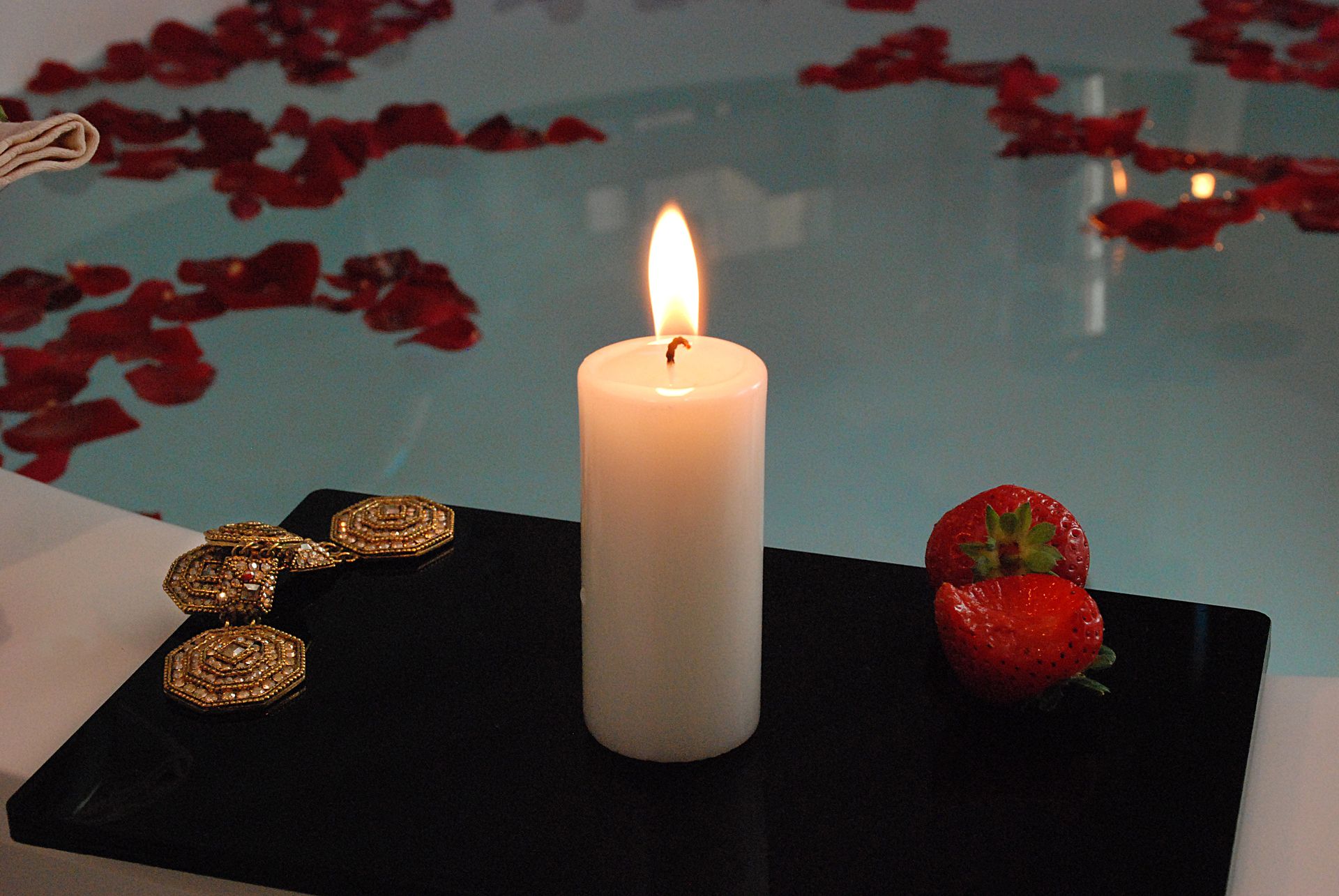 На черном подносе рядом с клубникой стоит зажженная белая свеча, а позади нее в воде в ванне плавают лепестки роз.