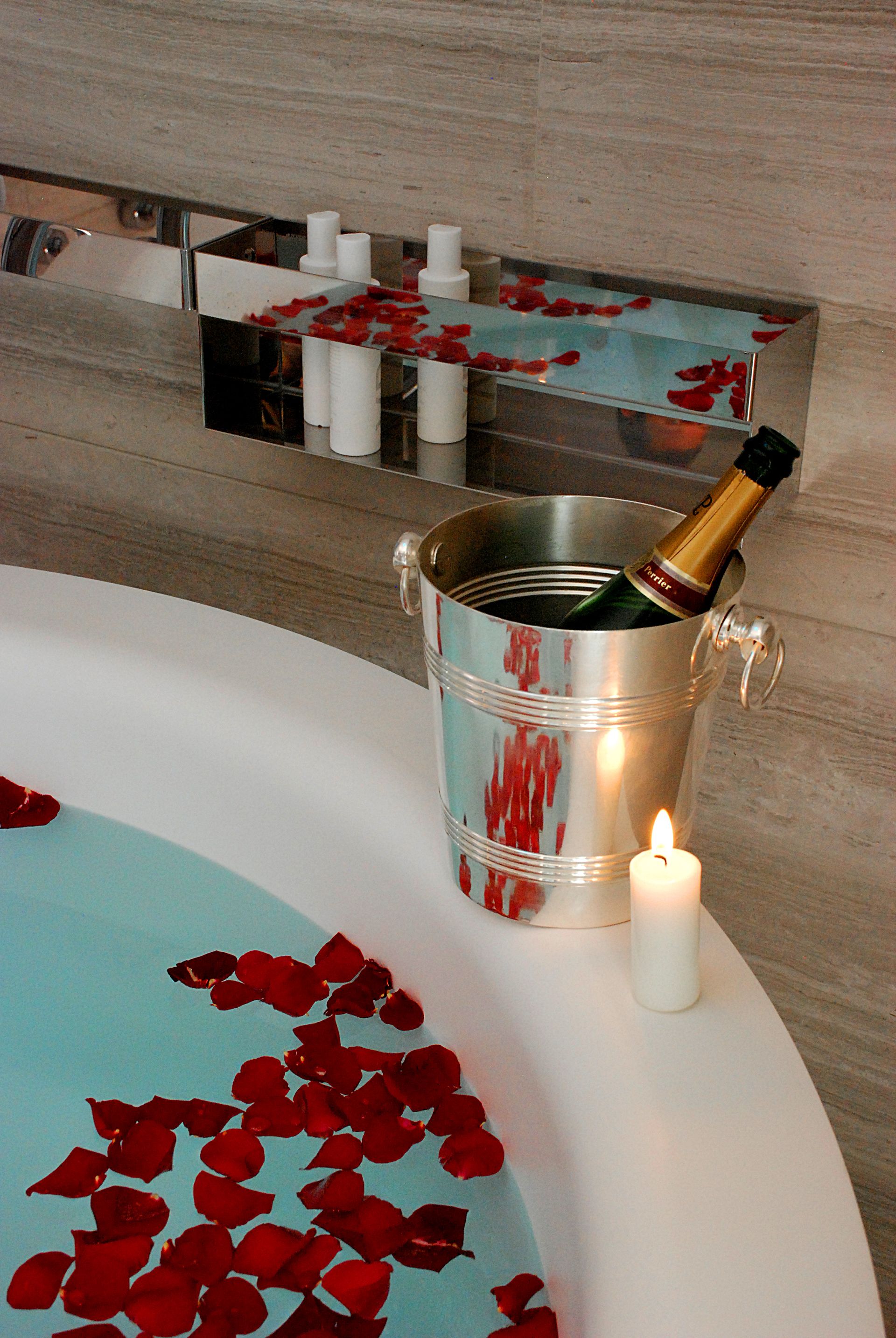 浴缸里盛满了蓝色的水和红色的玫瑰花瓣，浴缸边上放着一个香槟桶和一支点燃的蜡烛。