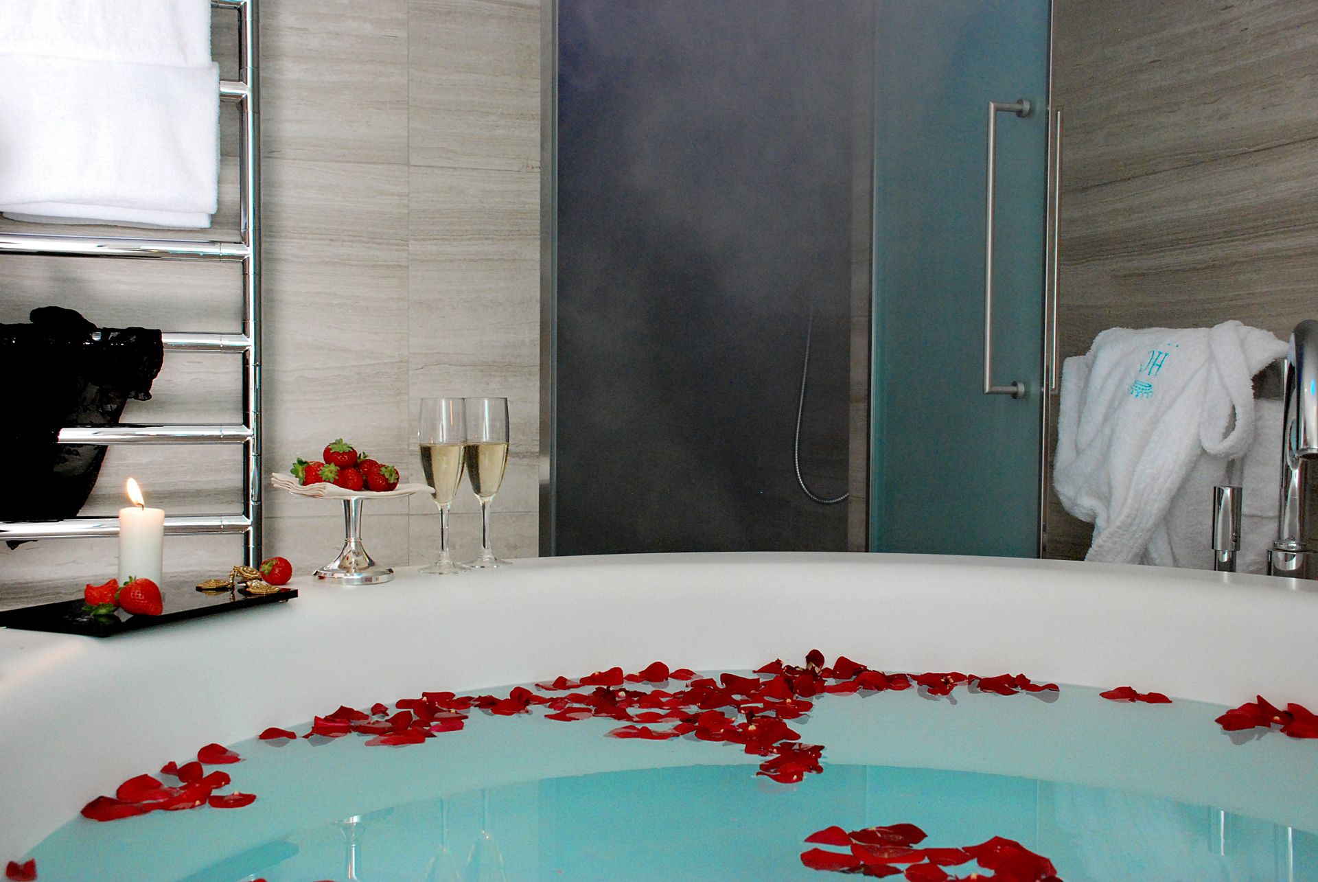 В ванной комнате — ванна, наполненная водой и лепестками красных роз, рядом бокалы с шампанским и клубника.