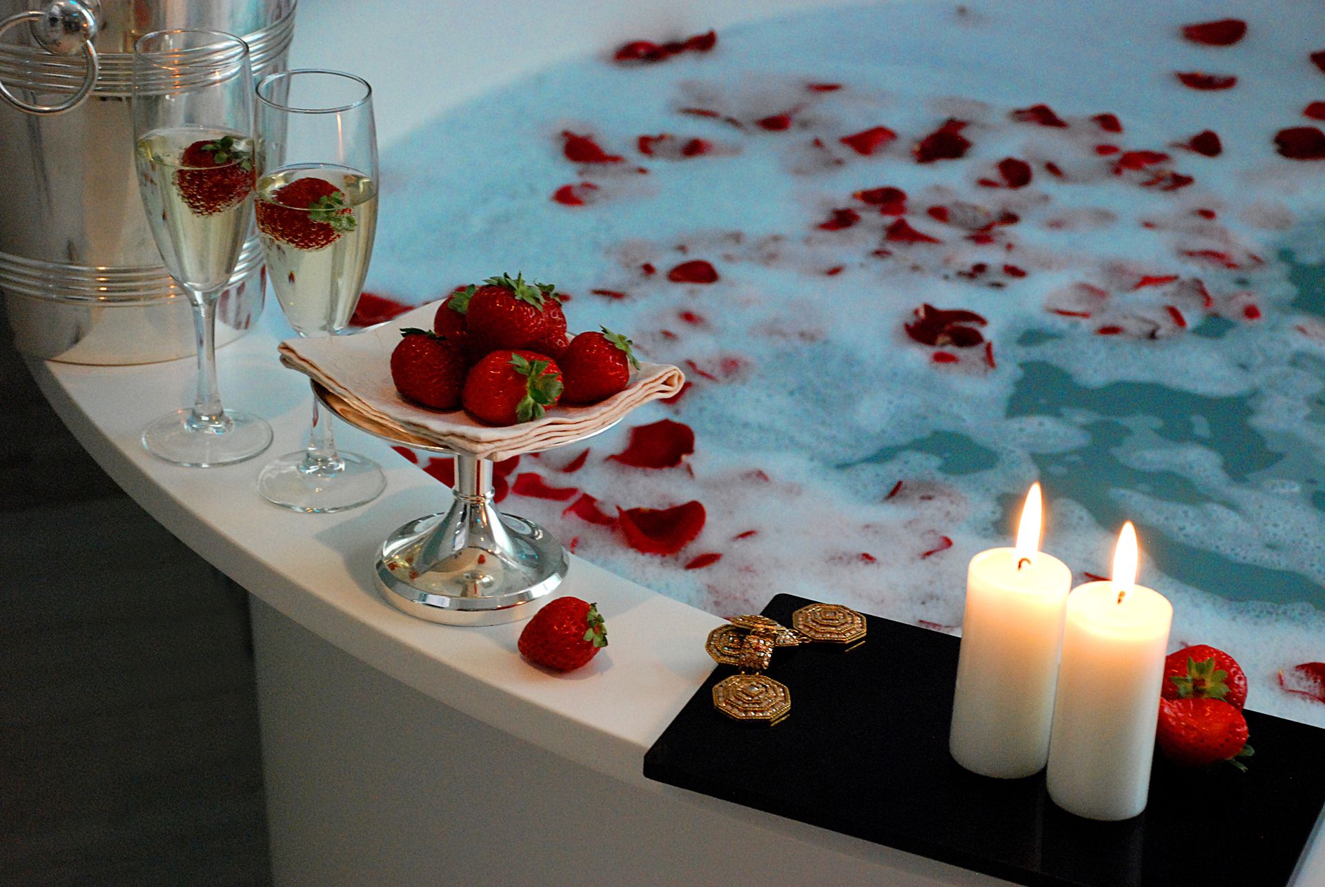 Ванна, освещенная свечами, с пеной и лепестками роз, бокалами для шампанского и тарелкой клубники на краю.