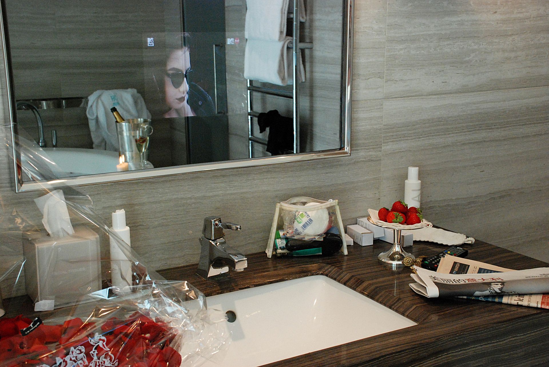 Туалетный столик в ванной комнате с белой раковиной, коробкой для салфеток, небольшой миской клубники и зеркалом, в котором отражается человек.