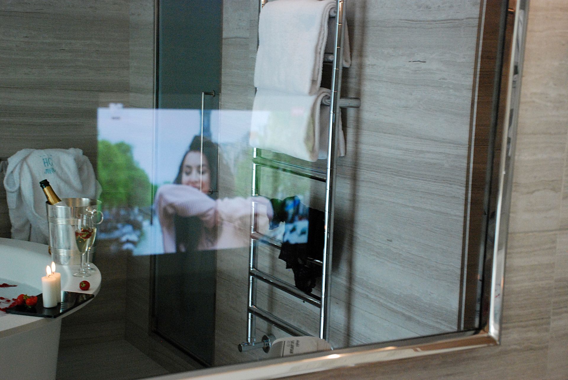 Зеркало в ванной комнате со встроенным телевизионным экраном, на котором изображен человек, рядом с ванной, шампанским и свечами.