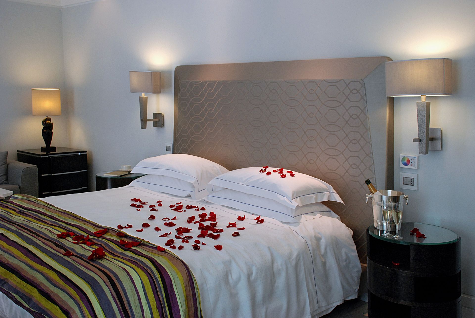 Романтическая гостиничная кровать с белым постельным бельем, разбросанными лепестками красных роз, полосатым пледом и шампанским в серебряном ведерке.
