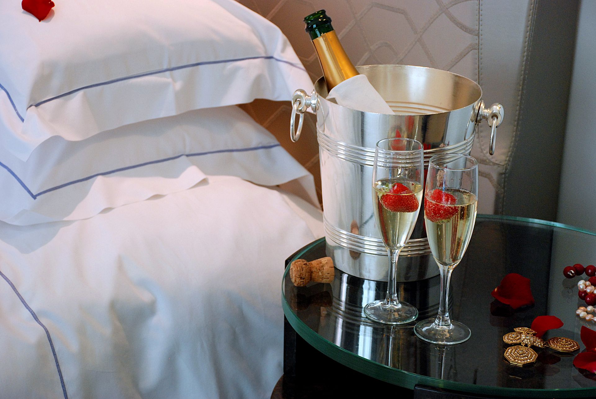 Бутылка шампанского в серебряном ведерке для льда и два бокала с лепестками роз, расставленные на прикроватной тумбочке с белыми подушками.