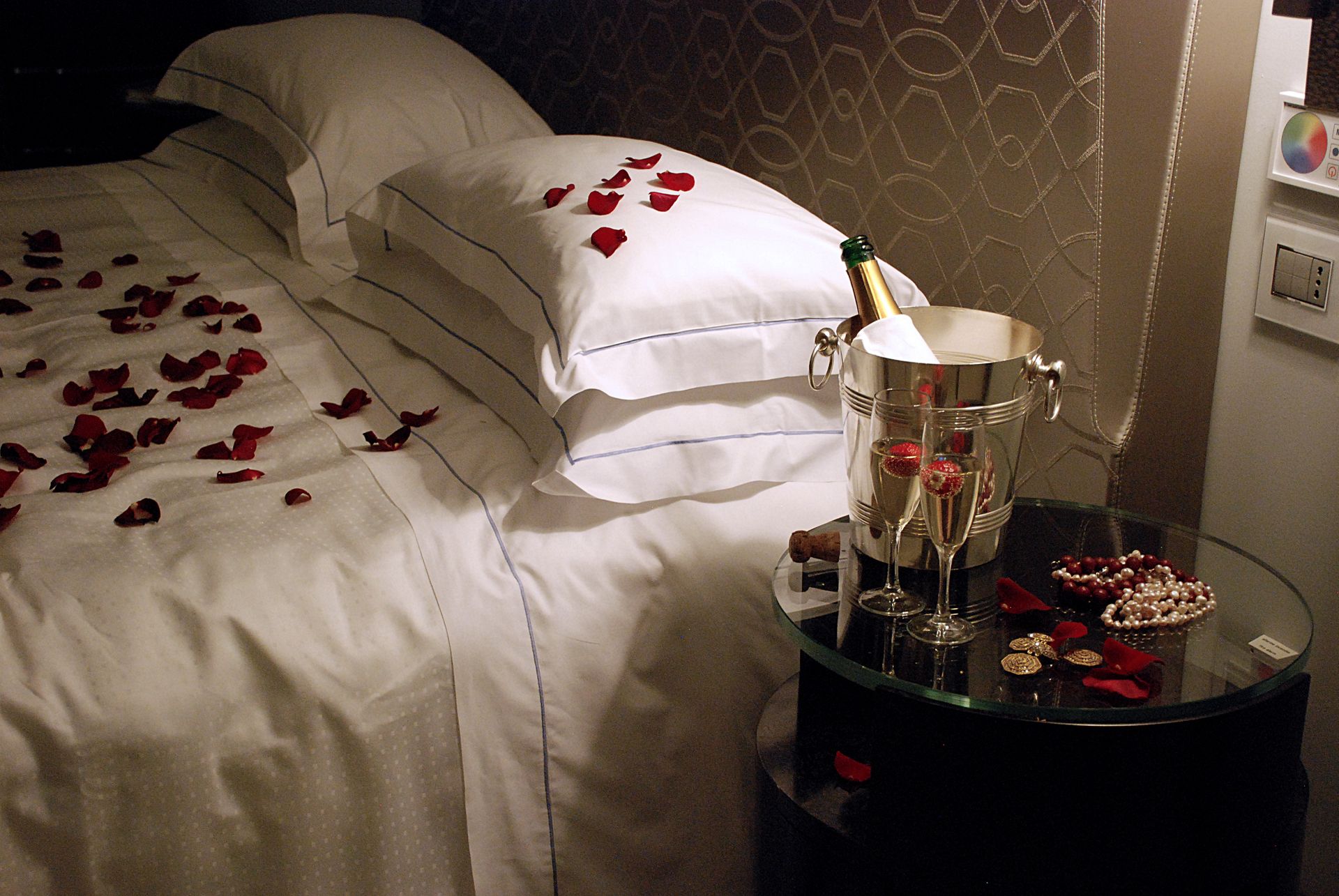 Кровать, покрытая лепестками роз на белых простынях, и тумбочка с ведерком для шампанского и двумя бокалами в полумраке комнаты.