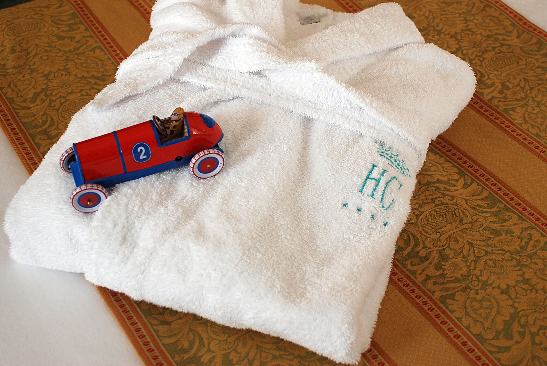 一件白色毛绒浴袍，上面印有字母“HC”字样，置于图案面料上，上面放着一辆红色玩具赛车。