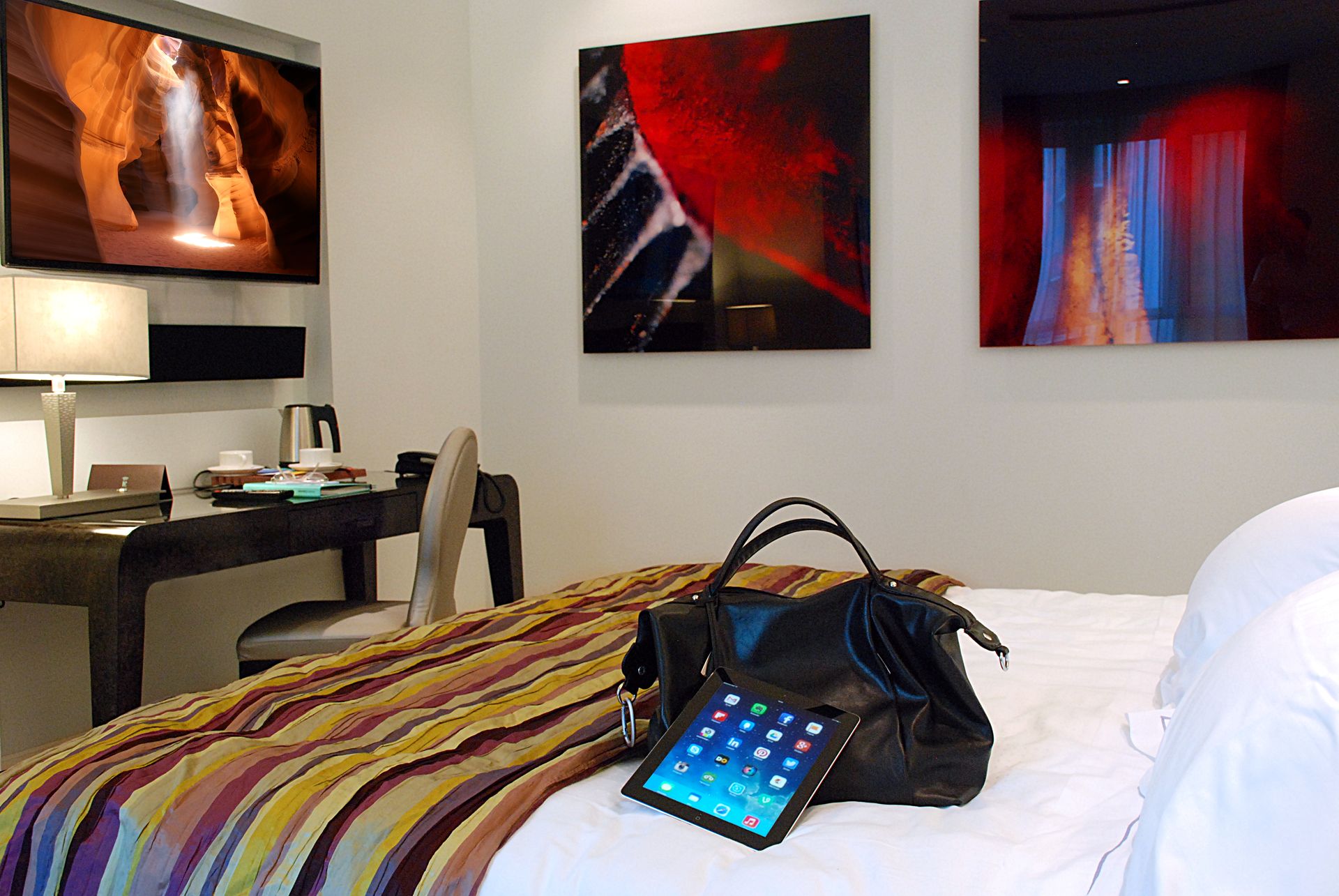 В гостиничном номере, на кровати, покрытой полосатым одеялом, лежат черная сумочка и планшет. На стенах также есть картины и письменный стол.