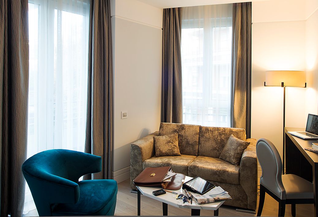 Гостиничный номер с бирюзовым креслом, диваном с узором, журнальным столиком с вещами и письменным столом с лампой и ноутбуком.