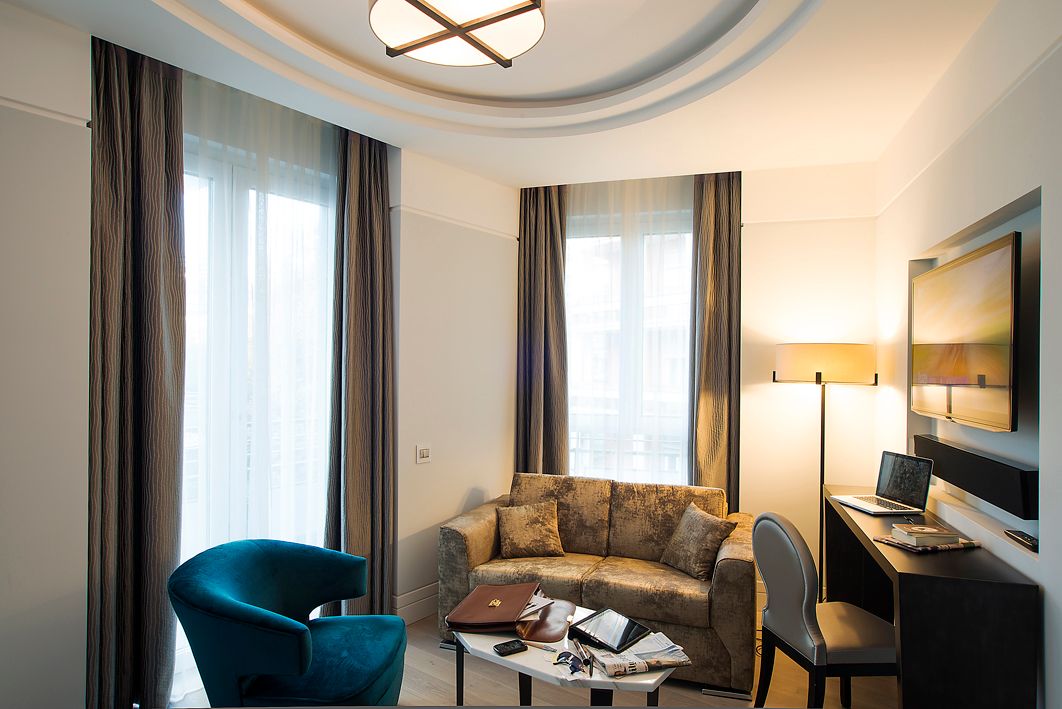 Гостиничный номер с бирюзовым креслом, диваном с узором, небольшим журнальным столиком, письменным столом, лампой и настенным телевизором.