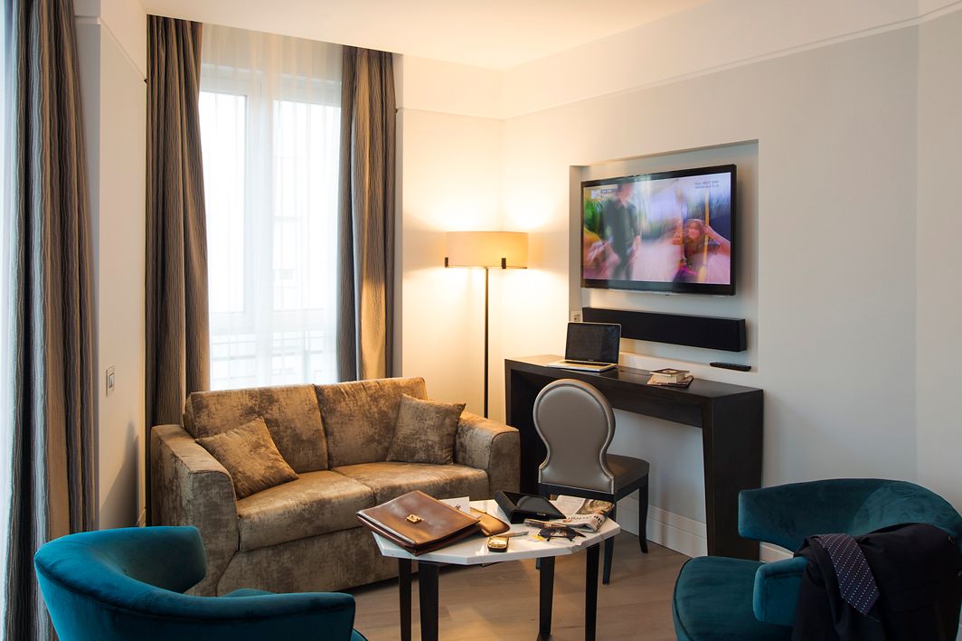 Гостиная зона с диваном бежевого цвета, двумя бирюзовыми креслами, журнальным столиком, письменным столом со стулом и настенным телевизором.