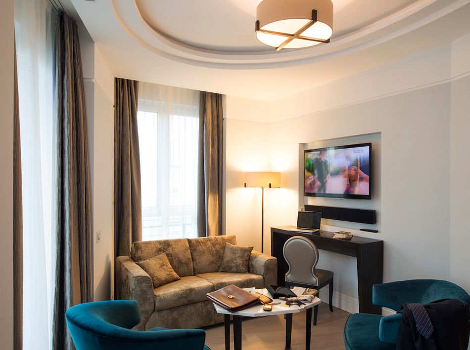 Гостиничный номер с бежевым диваном, двумя бирюзовыми креслами, журнальным столиком и письменным столом с настенным телевизором.