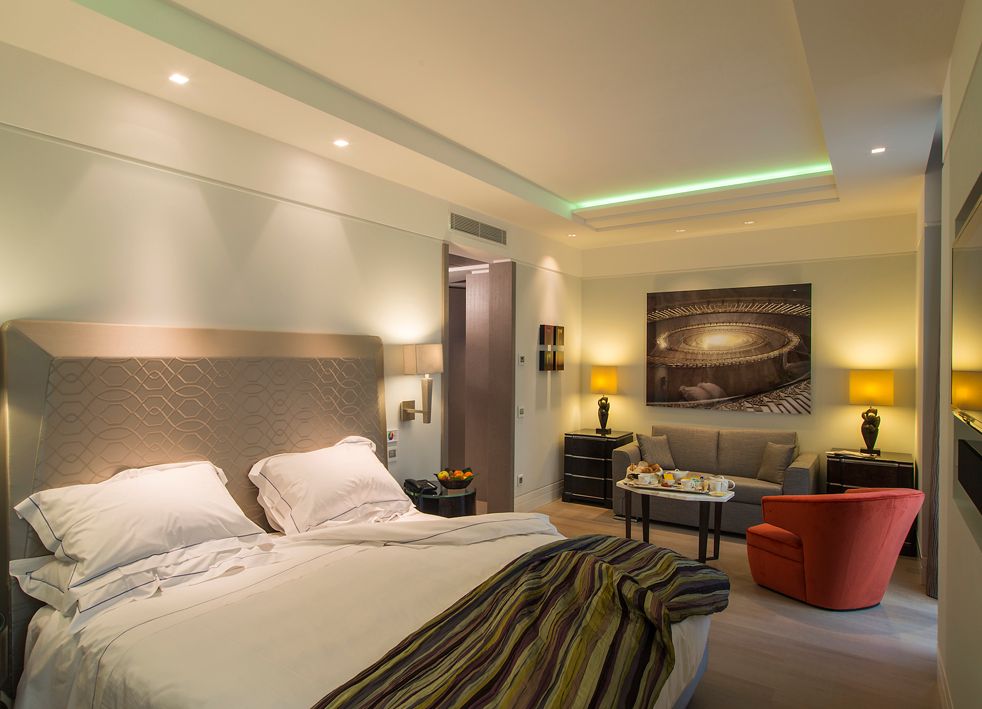 Современный гостиничный номер с большой кроватью, зоной отдыха, включающей серый диван и оранжевое кресло, а также теплым акцентным освещением.