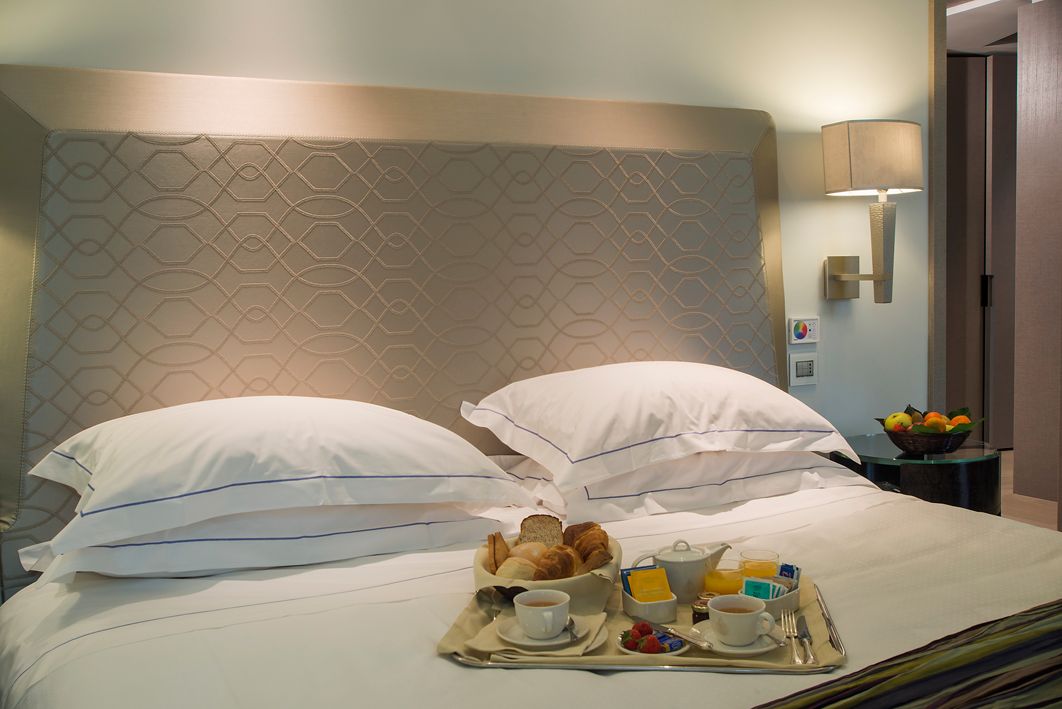 На белой кровати в гостиничном номере с фактурным изголовьем и настенным светильником стоит поднос с завтраком, выпечкой и кофе.