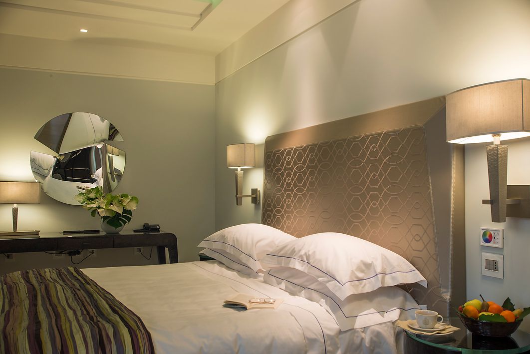 Номер в отеле с двуспальной кроватью, фактурным изголовьем, прикроватными лампами, круглым зеркалом и столиком с фруктами.