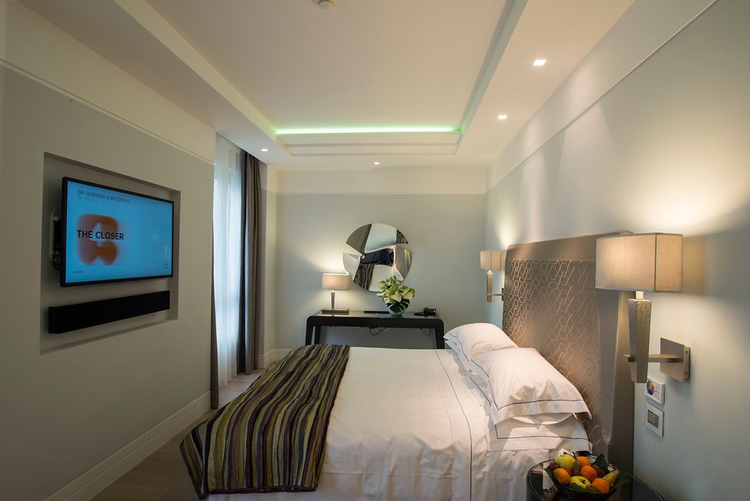 Современный, хорошо освещенный номер в отеле, оборудованный двуспальной кроватью, настенным телевизором и письменным столом с зеркалом и лампой.