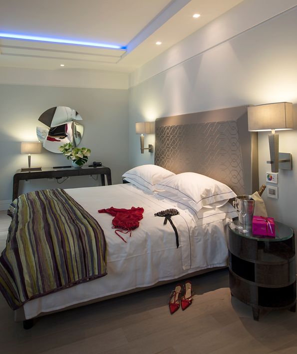 Современный гостиничный номер с кроватью, красной одеждой, черным аксессуаром и парой красных туфель на высоком каблуке на полу.