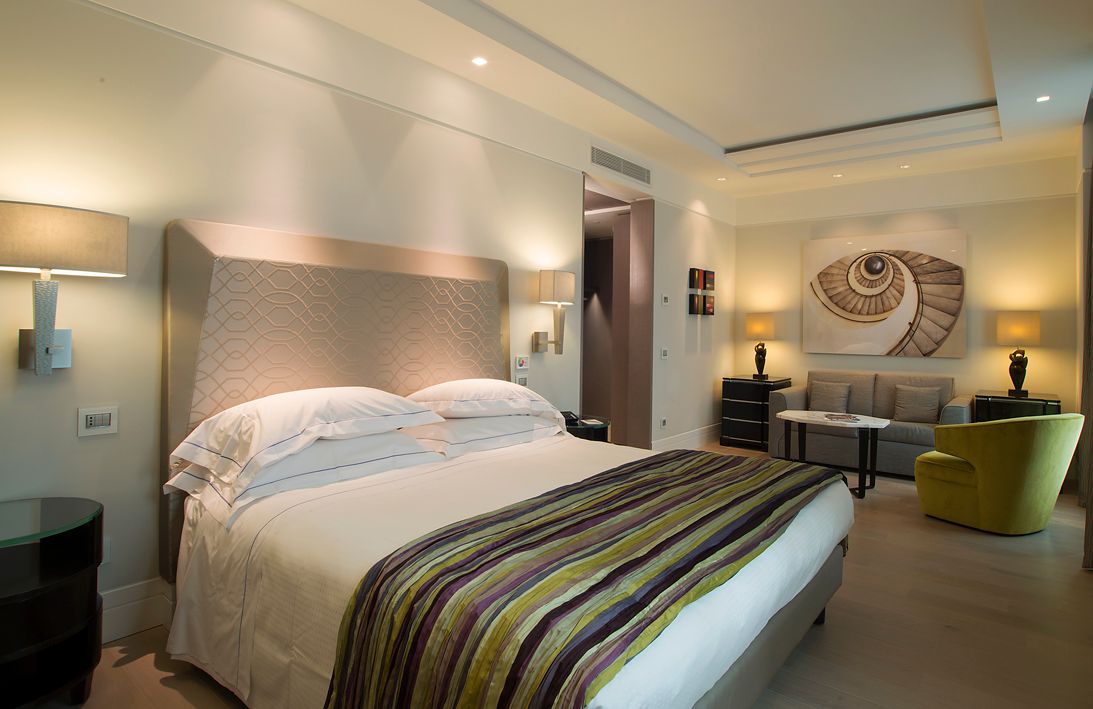 Современный гостиничный номер с большой кроватью, фактурным изголовьем, зоной отдыха с зеленым креслом и абстрактной картиной на стене.