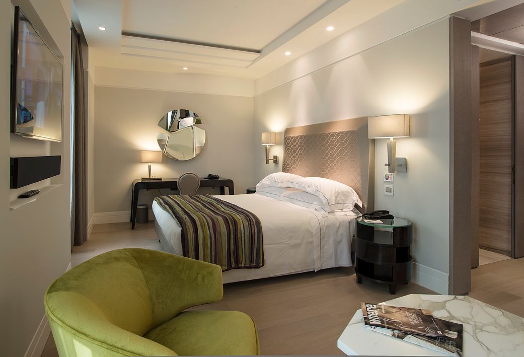 Современный гостиничный номер с кроватью, застеленной белым постельным бельем, зеленым креслом, мраморным столом и письменным столом с зеркалом.