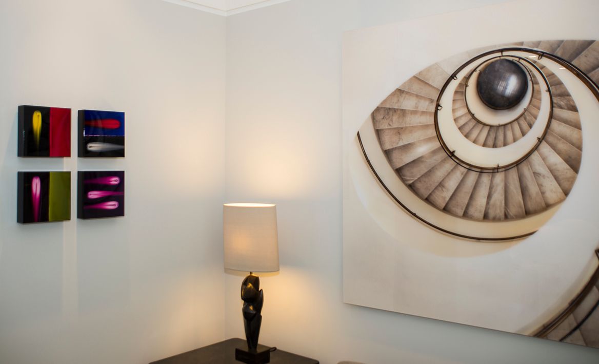 Между стеной, украшенной четырьмя небольшими квадратными панно в стиле абстрактных картин, и большим полотном с изображением винтовой лестницы стоит настольная лампа.