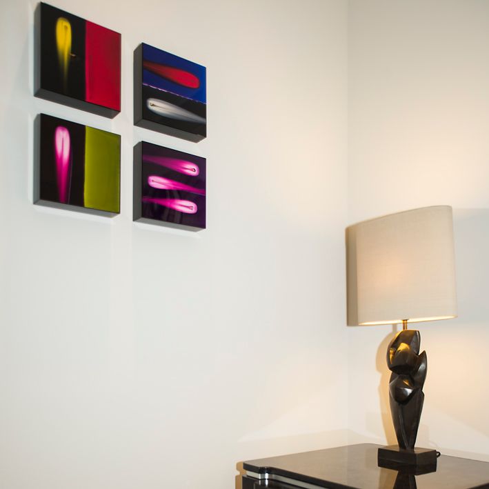 Над темной лампой на прикроватном столике на стене висят четыре абстрактные картины с изображением светящихся линий.