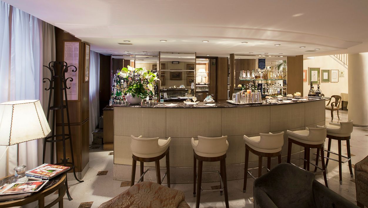 Изогнутая барная стойка с четырьмя бежевыми стульями в мягко освещенном современном помещении, слева от которой расположены лампа и полка.