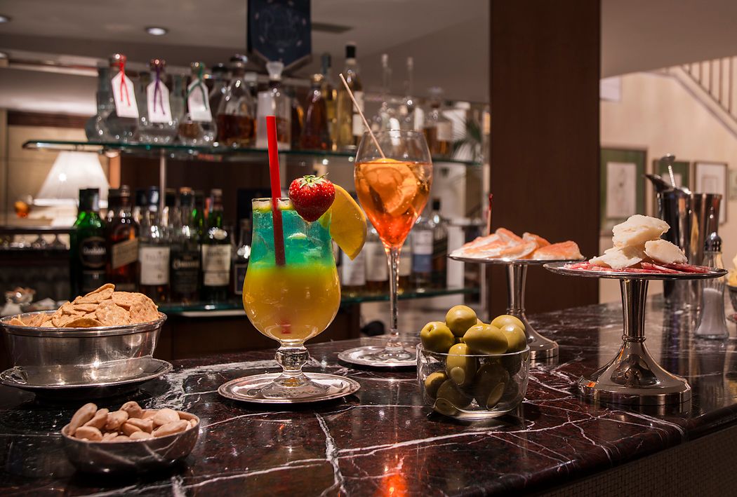 На темной барной стойке с мраморной столешницей перед полками расставлены два ярких коктейля, миска с орехами и закуски.