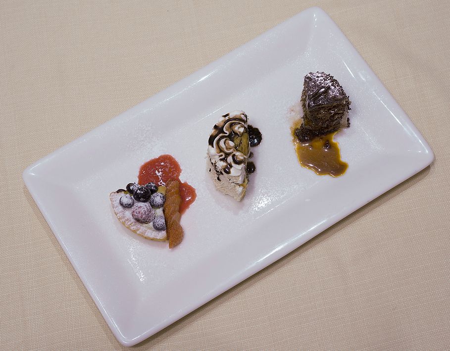 На белой прямоугольной тарелке представлены три небольших изысканных десерта с фруктами, сливками и шоколадным соусом.